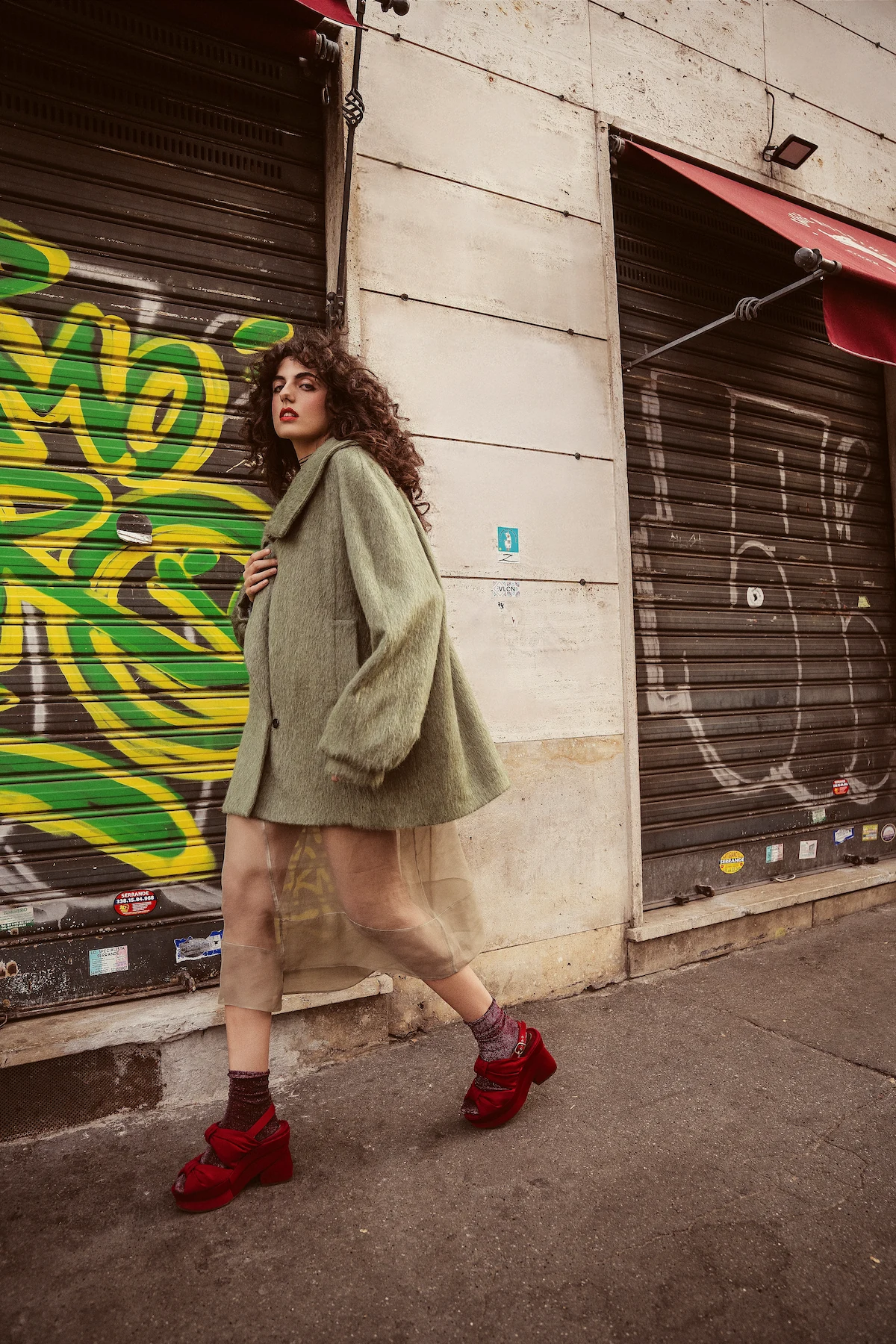 Look con cappotto verde salvia, gonna leggera e scarpe rosse davanti a una serranda urbana